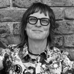 Headshot of Paul Montgomery, Unique Outcomes, wearing a floral shirt leaning against a brick wall