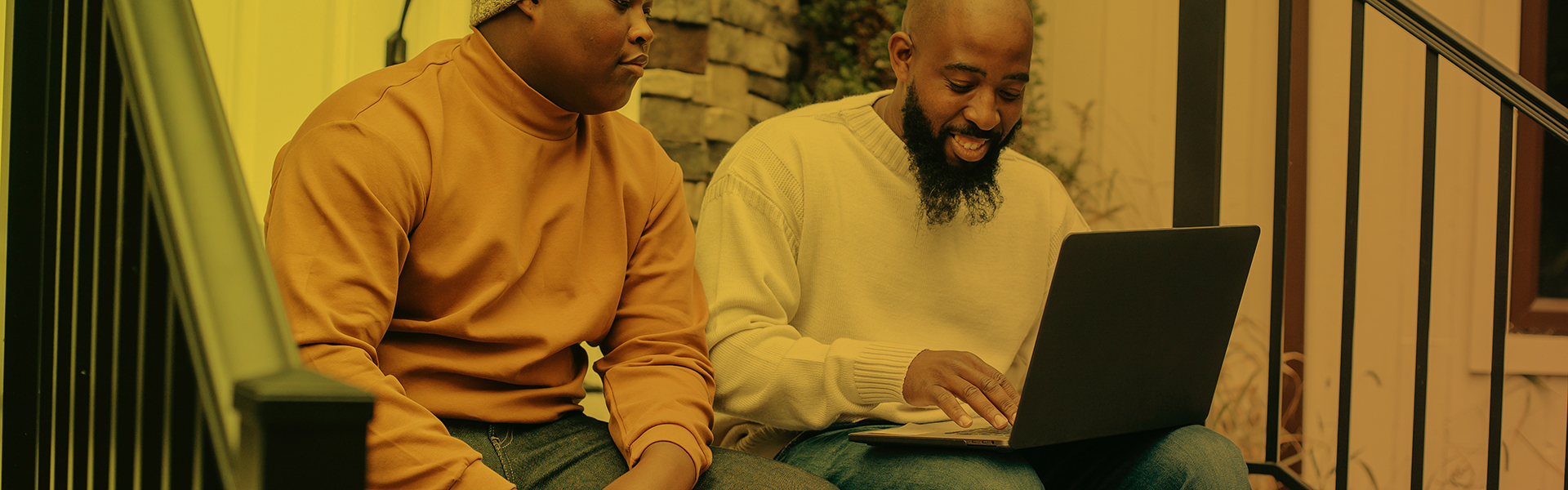 Stock image showing two people, both men, sitting on a staircase, looking at a laptop and smiling