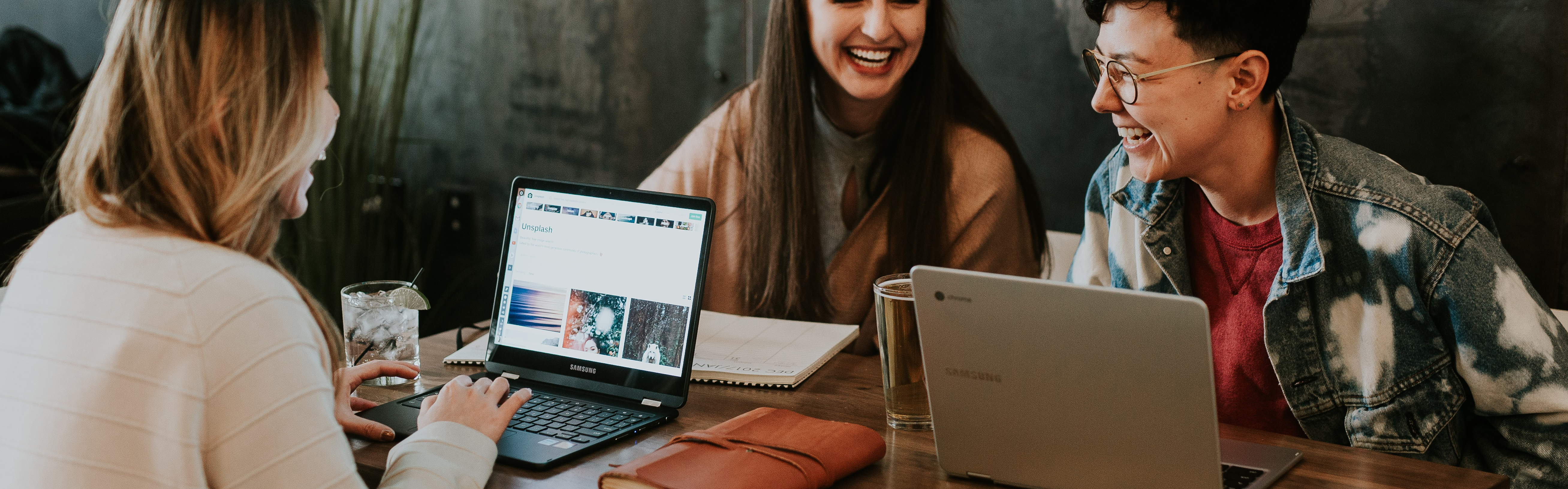 Stock cropped image showing 3 people sitting at a table, with laptops, talking and laughing