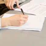 A cropped photographc showing 2 people in black and white blouses sitting at a large beige table, looking at pieces of paper, one is holding a pen as if taking notes