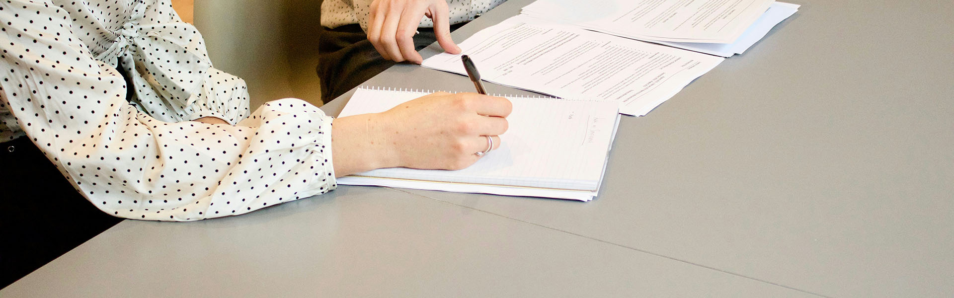 A cropped photographc showing 2 people in black and white blouses sitting at a large beige table, looking at pieces of paper, one is holding a pen as if taking notes