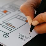 A cropped close up image showing a piece of paper with several frame boxes and hand holding a pen above the words User Journey.