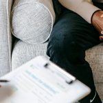 Cropped image showing a person sitting on a couch with clasped hands, and another person sitting opposite them holding a clipboard and a pen