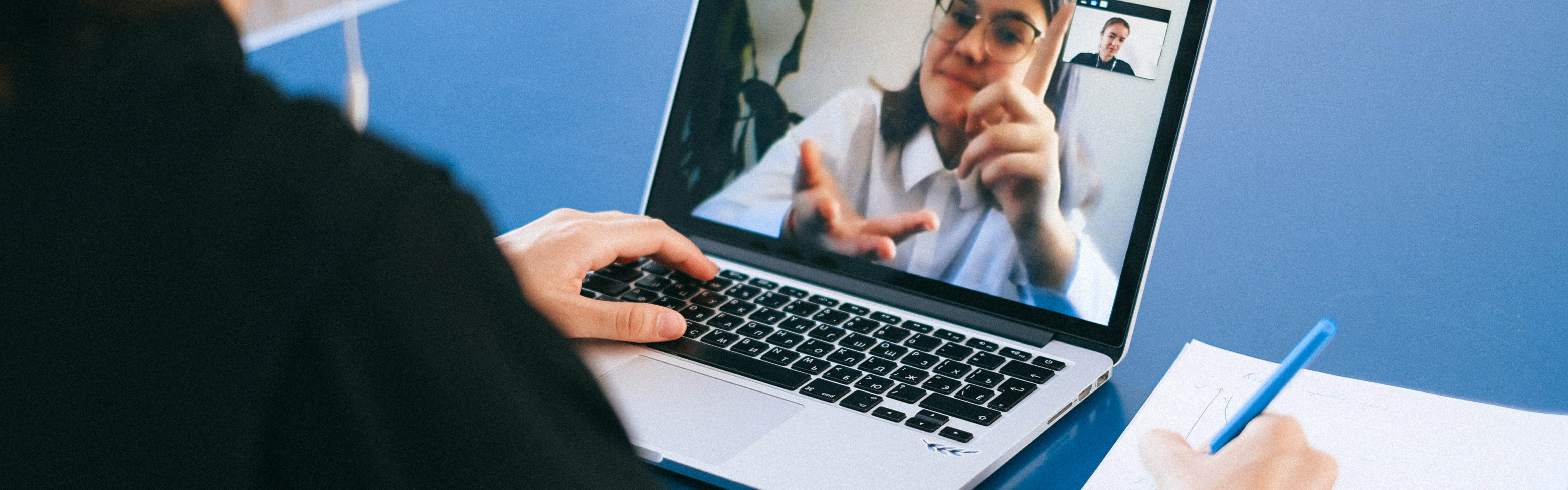 Stock image looking over someones shoulder while writing on a notebook, looking at a laptop screen showing a woman gesturing and talking via zoom