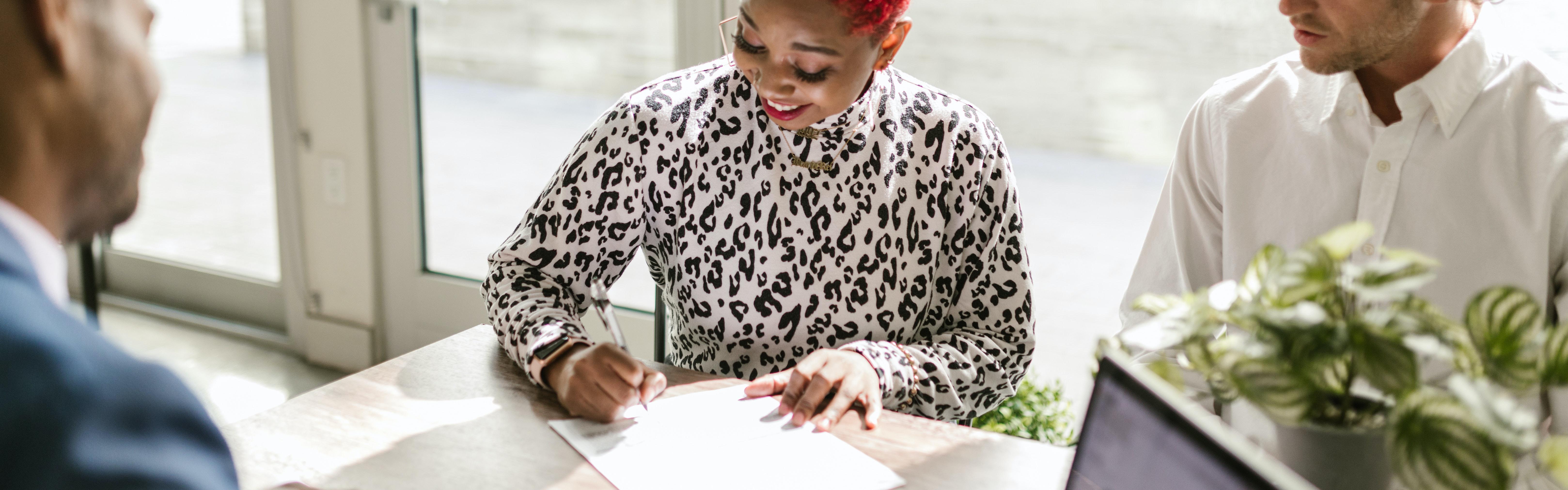 Stock photograph showing 3 people sitting at a desk, smiling while one writes notes.