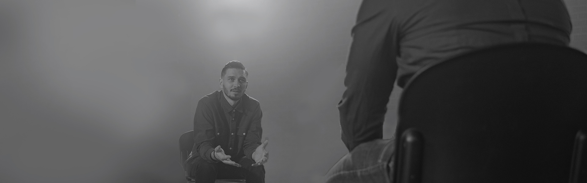 Stock image showing a man sitting on a chair gesturing and talking to another man (back only visible)